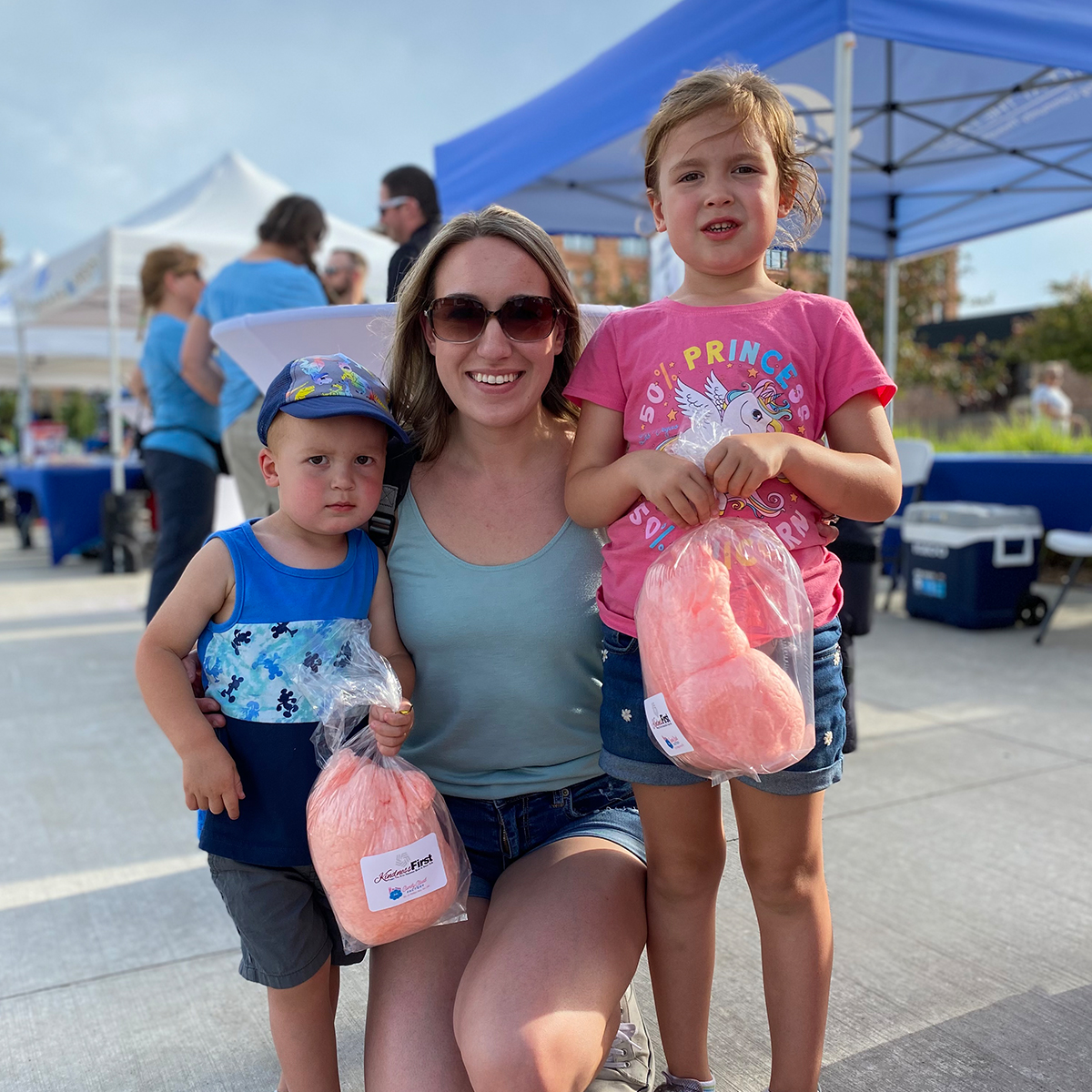 Mother kneels on ground with arms around her two children: an older daughter and younger son. Both children hold a bag of bright pink cotton candy.