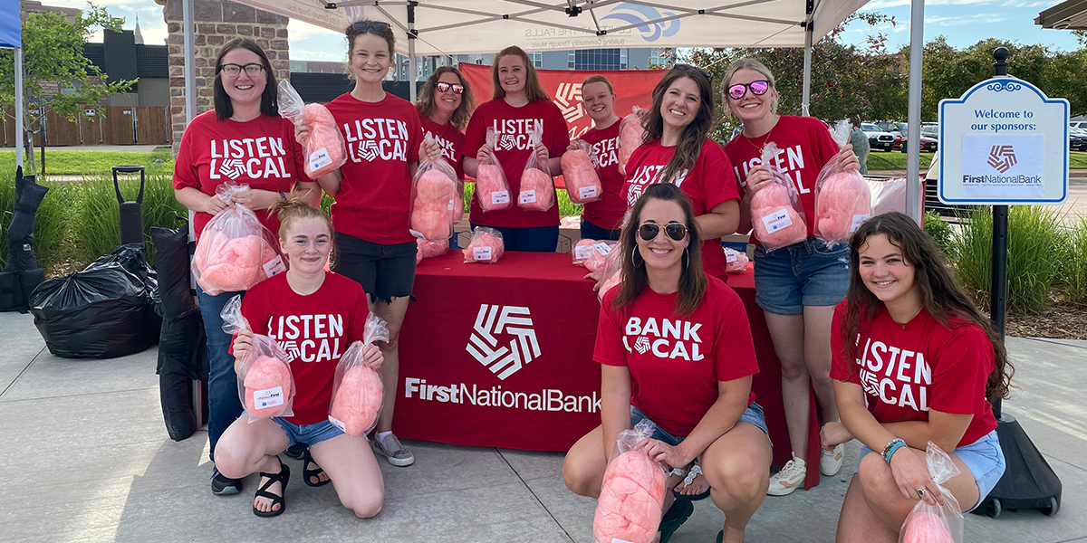 First National Bank employees wearing red shirts pose around booth, holding bags of bright pink cotton candy.