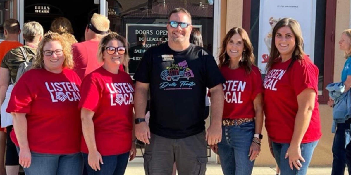 First National Bank employees pose with owner of Dells Theatre. A line of people stand in the background, waiting for free movie tickets.