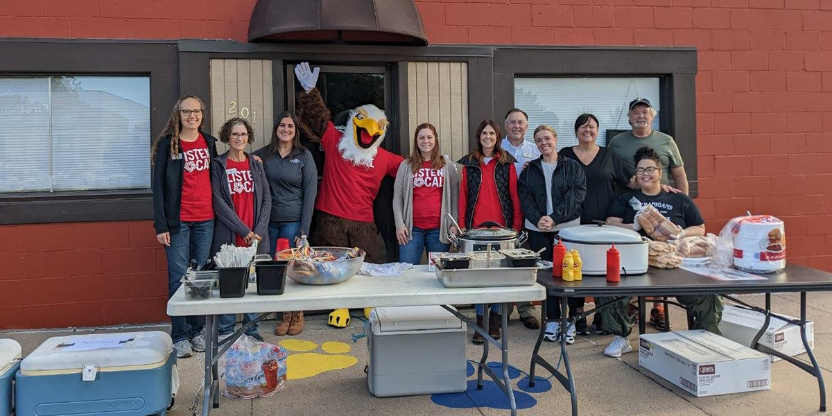 Group of First National Bank employees pose outside with Someday Café employees. In front are tables with hot dogs, buns, and condiments to serve.
