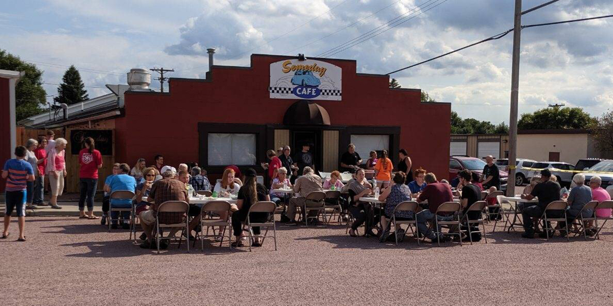 Group of First National Bank employees pose outside with Someday Café employees. In front are tables with hot dogs, buns, and condiments to serve.