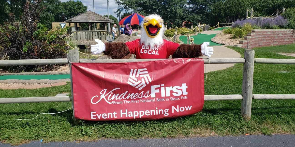 Eagle mascot wearing First National Bank shirt poses in front of banner that reads, "KindnessFirst from The First National Bank in Sioux Falls. Event happening now."
