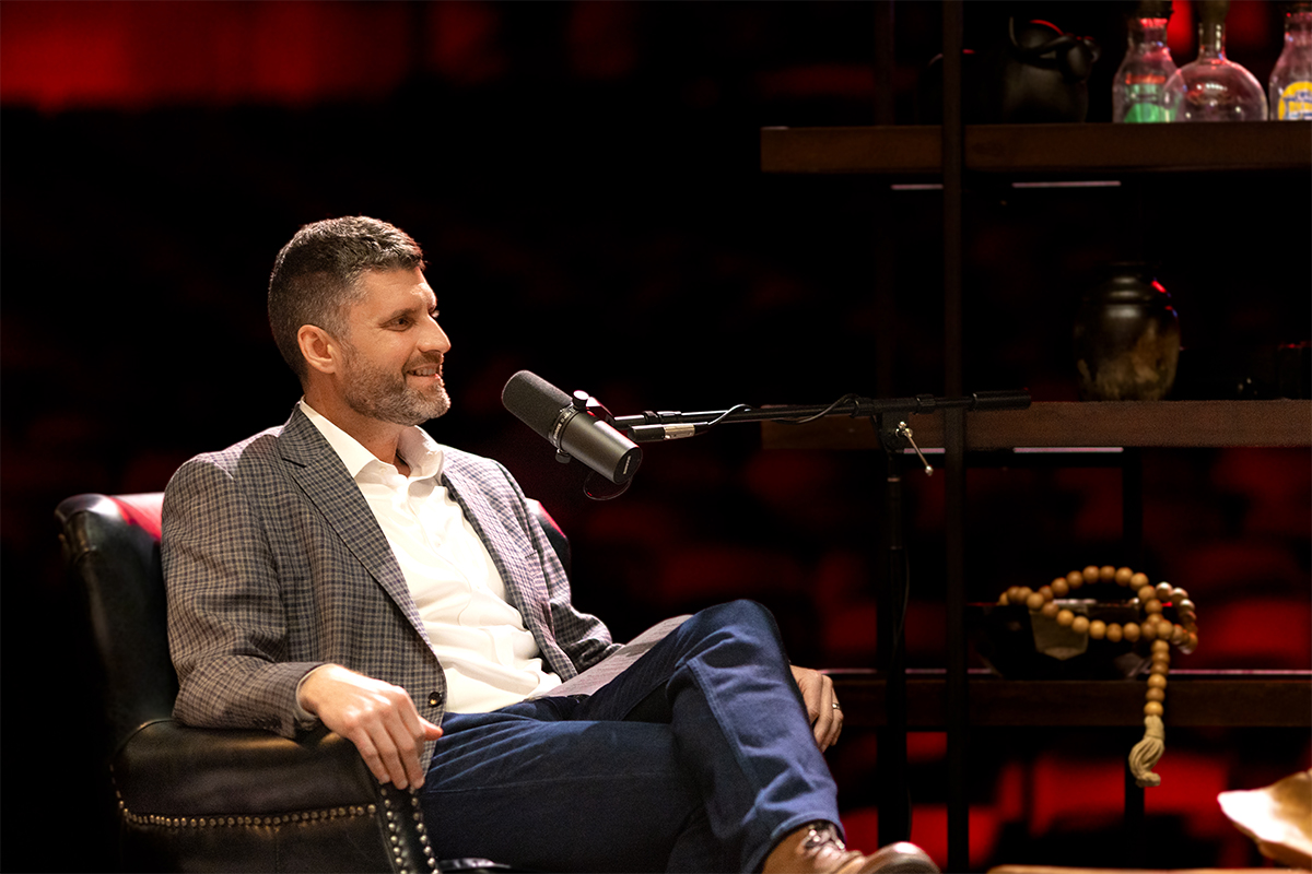 Adam Cox reclining in a leather chair, speaking into a microphone at the live Common Cents on the Prairie event.