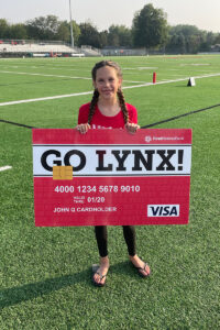 A young girl holds a large debt card that says "Go Lynx" while standing on a football field.