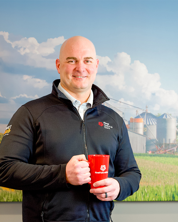 Phil DeGroot standing in front of a picture of a farm, holding a First National Bank mug.