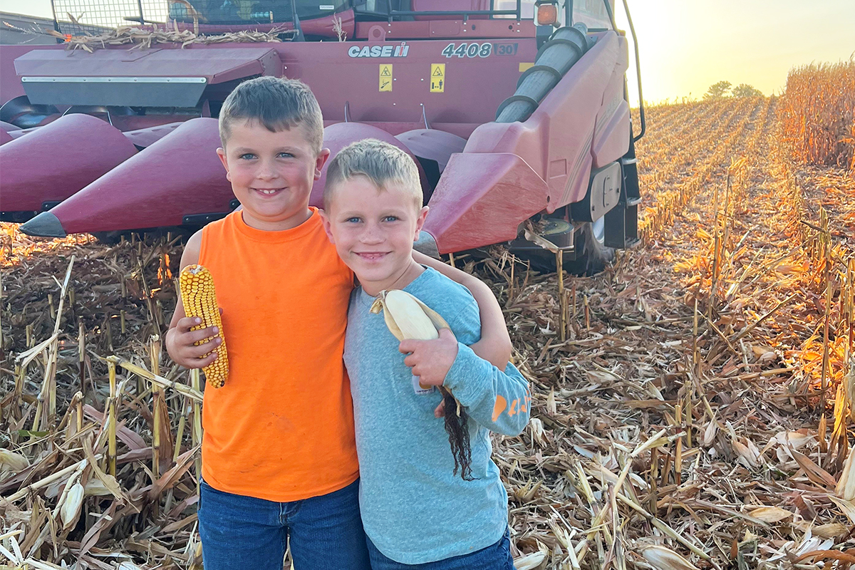 Two young boys standing in front of a combine in a corn field.