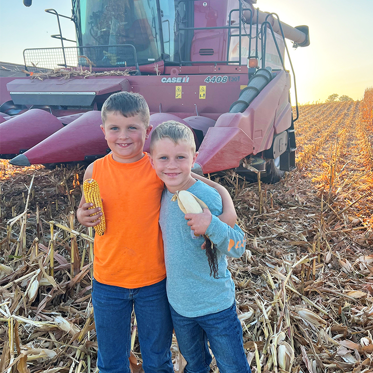 Two young boys standing in front of a combine in a corn field.