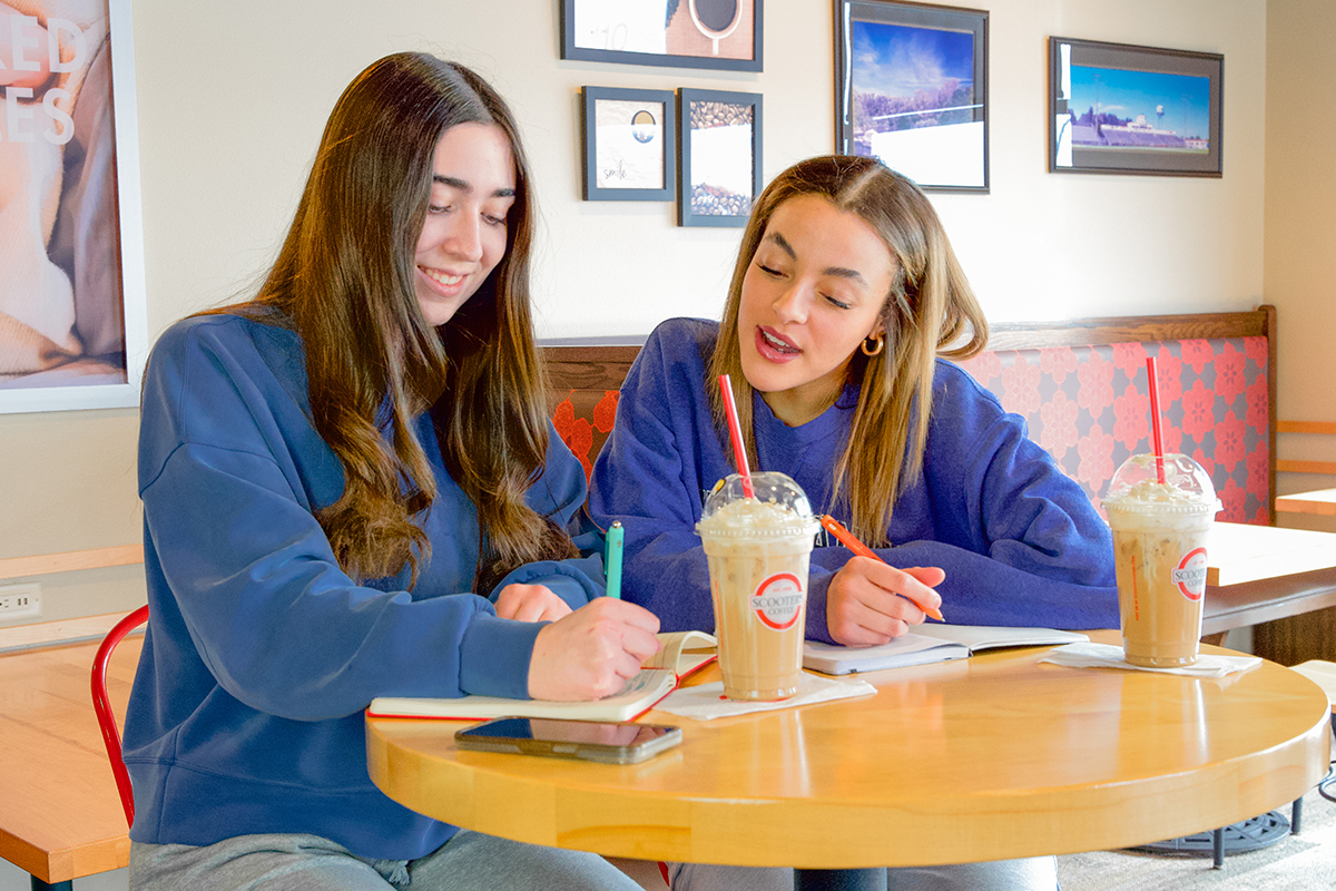 Two teenage girls writing in notebooks while sitting in a coffee shop.