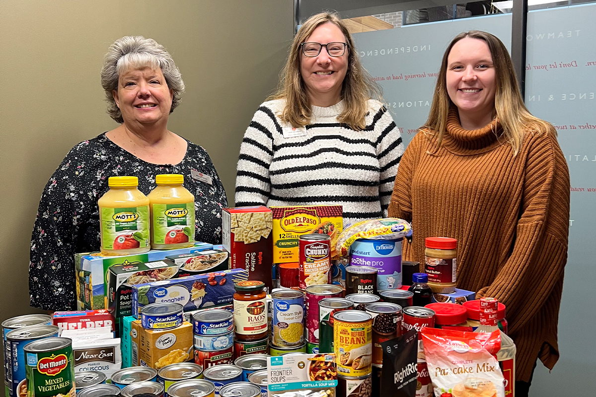 Three women with a stack of food items collected from a food drive.