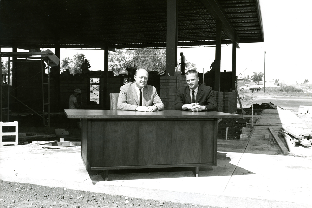 A photo from the 1960s showing two men sitting behind a desk with the frame of an unconstructed building in the background.