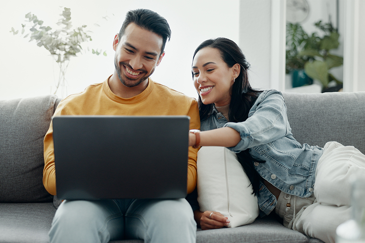 A young married couple sitting on a couch looking at their laptop screen.