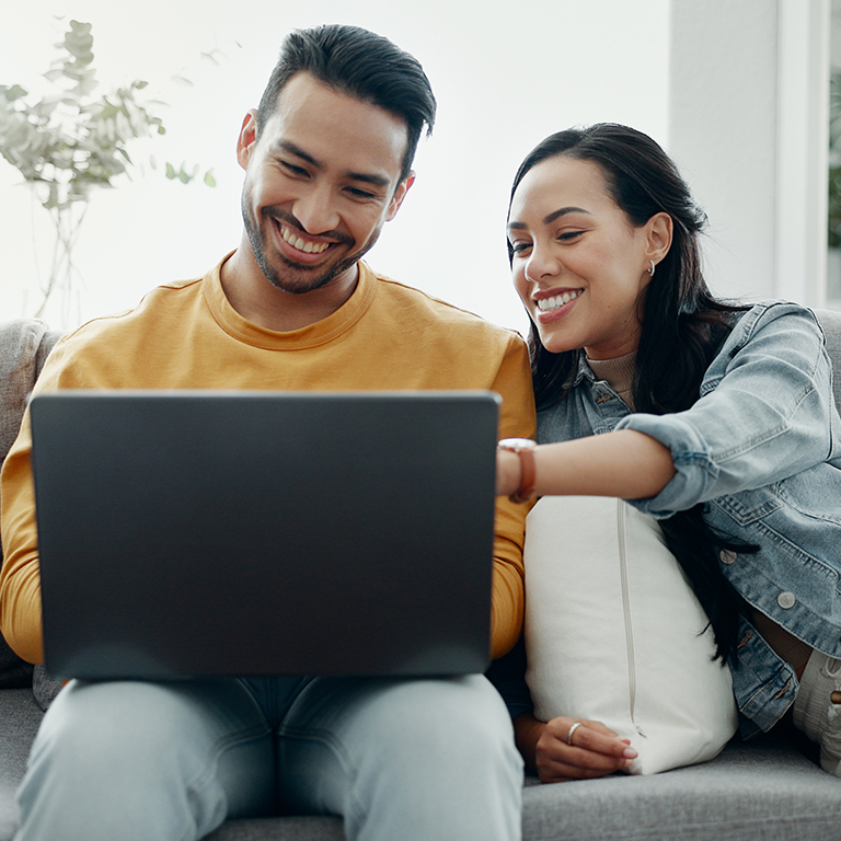 A young married couple sitting on a couch looking at their laptop screen.